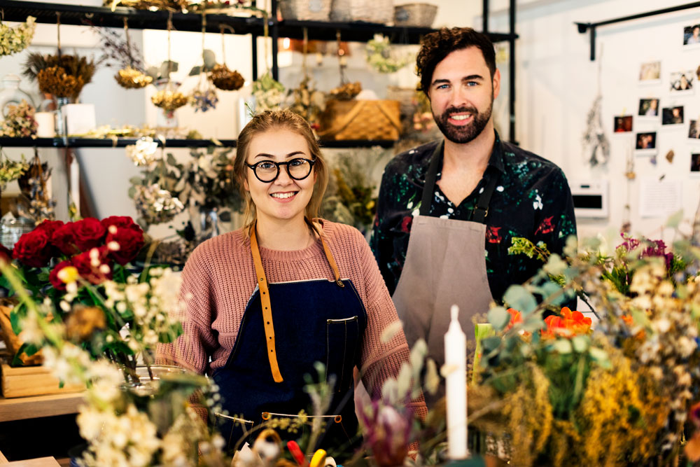 Flower shop owners smiling behind a display of fresh flowers and greenery, showcasing local business spirit in a vibrant floral setting.