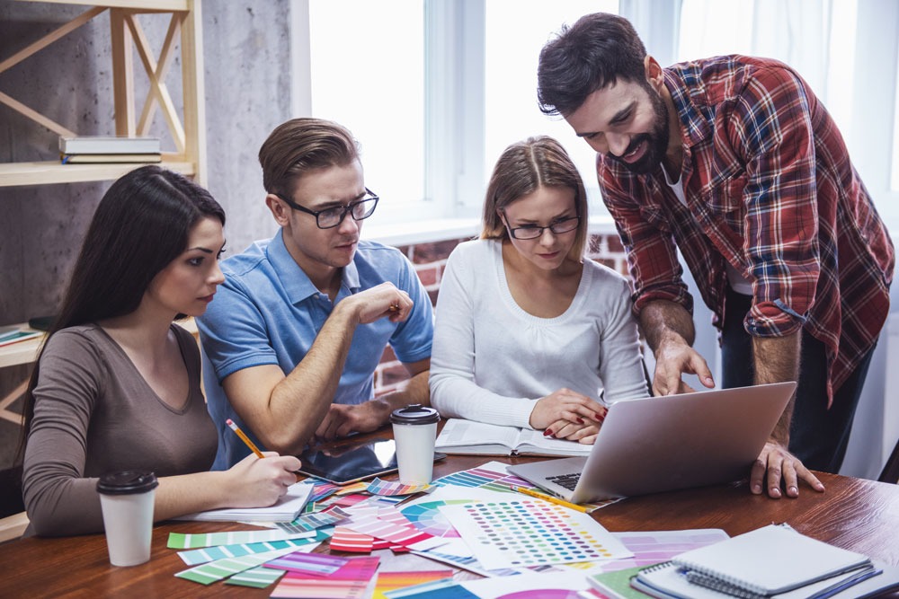 Group of four professionals collaborating around a laptop, discussing digital marketing strategies, with design materials and coffee cups on the table, reflecting teamwork in SEO and digital marketing services.
