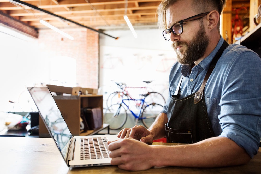 Man in apron using laptop in a creative workspace, with bicycles visible in the background, illustrating digital marketing and SEO services for local businesses.