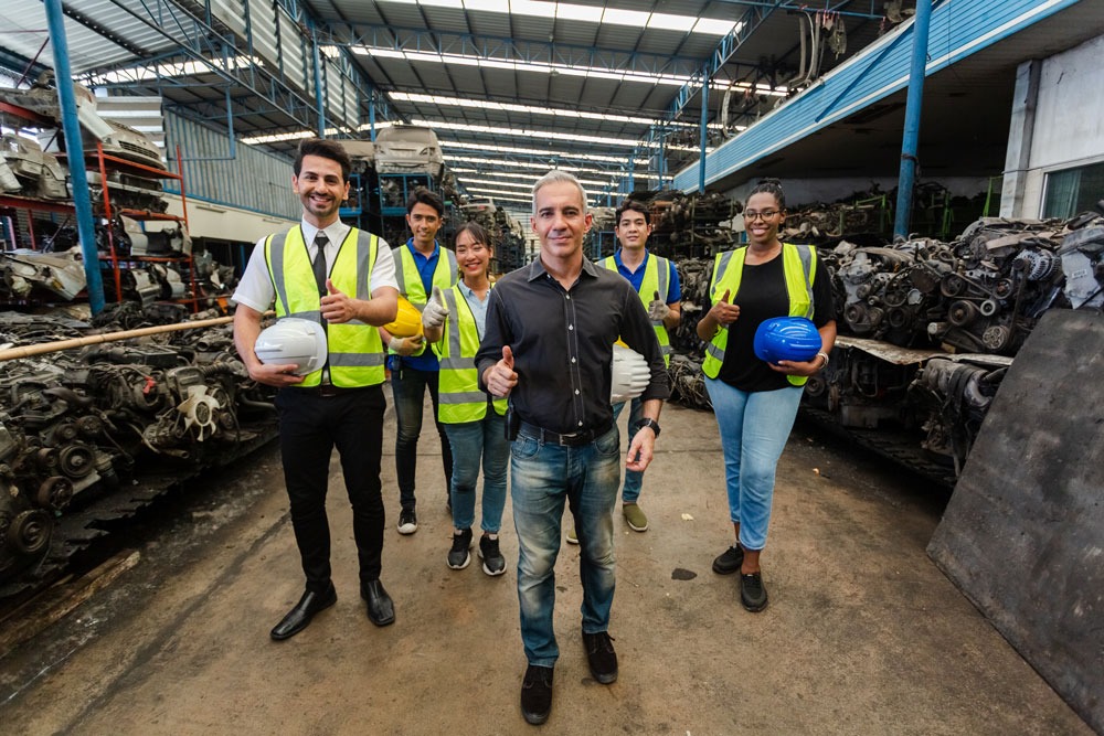 Group of diverse individuals in safety vests and helmets, standing confidently in an automotive workshop surrounded by car parts, representing teamwork and professionalism in the automotive industry.
