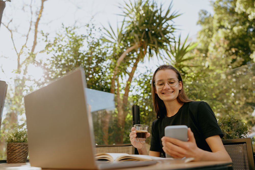 Young businesswoman smiling while reading a message on her smartphone, seated at a table with a laptop and a drink, surrounded by greenery, illustrating the modern work environment and digital engagement.