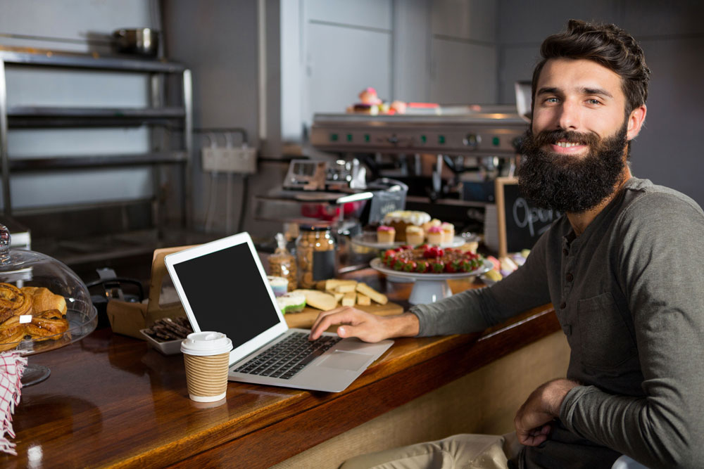 Bearded man typing on laptop and sipping coffee in a café, surrounded by pastries and desserts, illustrating the blend of digital marketing and local business engagement.