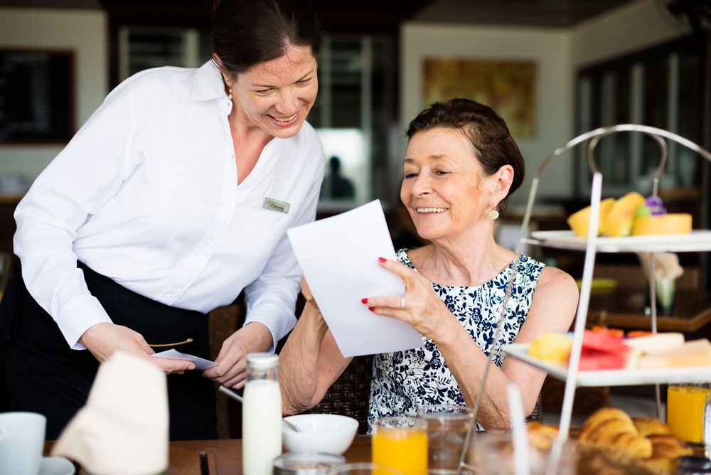 Waitstaff assisting a smiling older woman with a menu in a café setting, featuring a tiered stand of pastries and drinks, reflecting customer service and dining experiences.