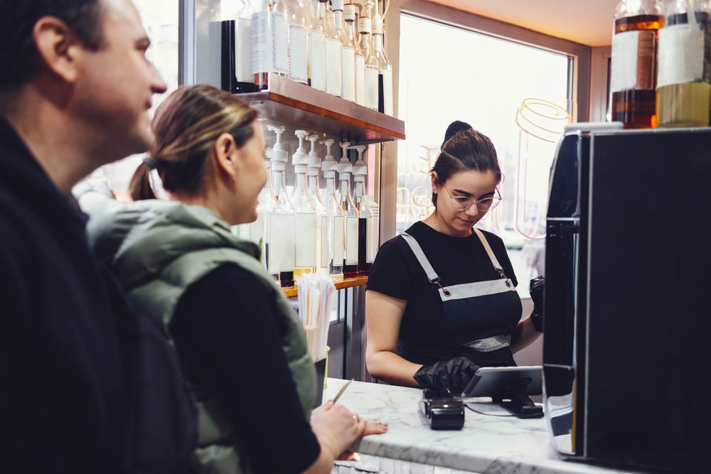 Barista serving customers at a café, with bottles of ingredients in the background, illustrating local business engagement and customer service in Wollongong.