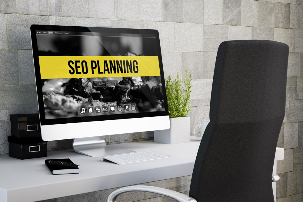 Desktop computer displaying "SEO PLANNING" on screen, with a modern office setup including a black chair, potted plant, and stationery, reflecting the digital marketing focus of an SEO agency.
